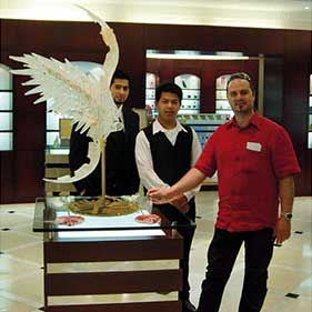 This is an image featuring three individuals standing in a showroom. The focal point of the image appears to be a large, intricate glass sculpture that resembles a bird with outstretched wings, positioned on a display stand. The man wearing a red shirt is gesturing towards the sculpture, possibly explaining its design or craftsmanship. Behind him and slightly to the side is another man in a dark-colored suit looking at the sculpture with interest, while a woman in a light-colored top stands behind both of them, also observing the artwork. The showroom itself has elegant lighting fixtures overhead and what looks like display cases that could be used for showcasing glassware or other delicate items. The setting suggests this might be an event or a store specializing in high-quality glass art, possibly in Venice, Italy, which is famous for its Murano glass craftsmanship. Keywords: MURANO GLASS, VASE, LAMP, MURANO GLASS, DESIGN, MADE IN ITALY, VENICE GLASS