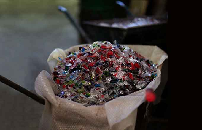 The image shows a bag of colorful glass shards or bits, which appear to be byproducts from the production process at a Murano glass factory. The colors are vibrant and varied, suggesting that these shards have been produced during the creation of different colored glass items. The background is dimly lit, which contrasts with the bright colors of the glass pieces, making them stand out in the image. There's also a glimpse of what looks like a workbench or table, indicating that this is likely an industrial setting where glassware is made. Keywords for this image might include: Murano glass, shards, byproducts, colorful glass, production waste, artisanal glass, Venice glass, and Italy glass-making.